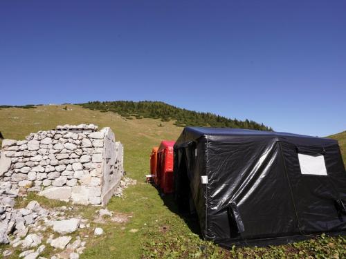En črn šotor, drugi rdeč in tretji oranžen stojijo pred ruševinami na planin nad Jesenicami.