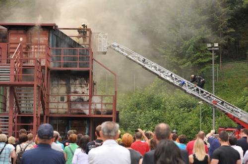 Gledalci spremljajo praktično vajo gašenja požara v objektu. Gasilec se vzpenja po lestvi, na stavbi k drugemu gasilcu.