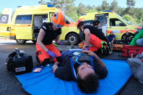 Dva reševalca izvajata triažo nad pacientom v ozadju sta dva reševalna vozila in tri osebe.